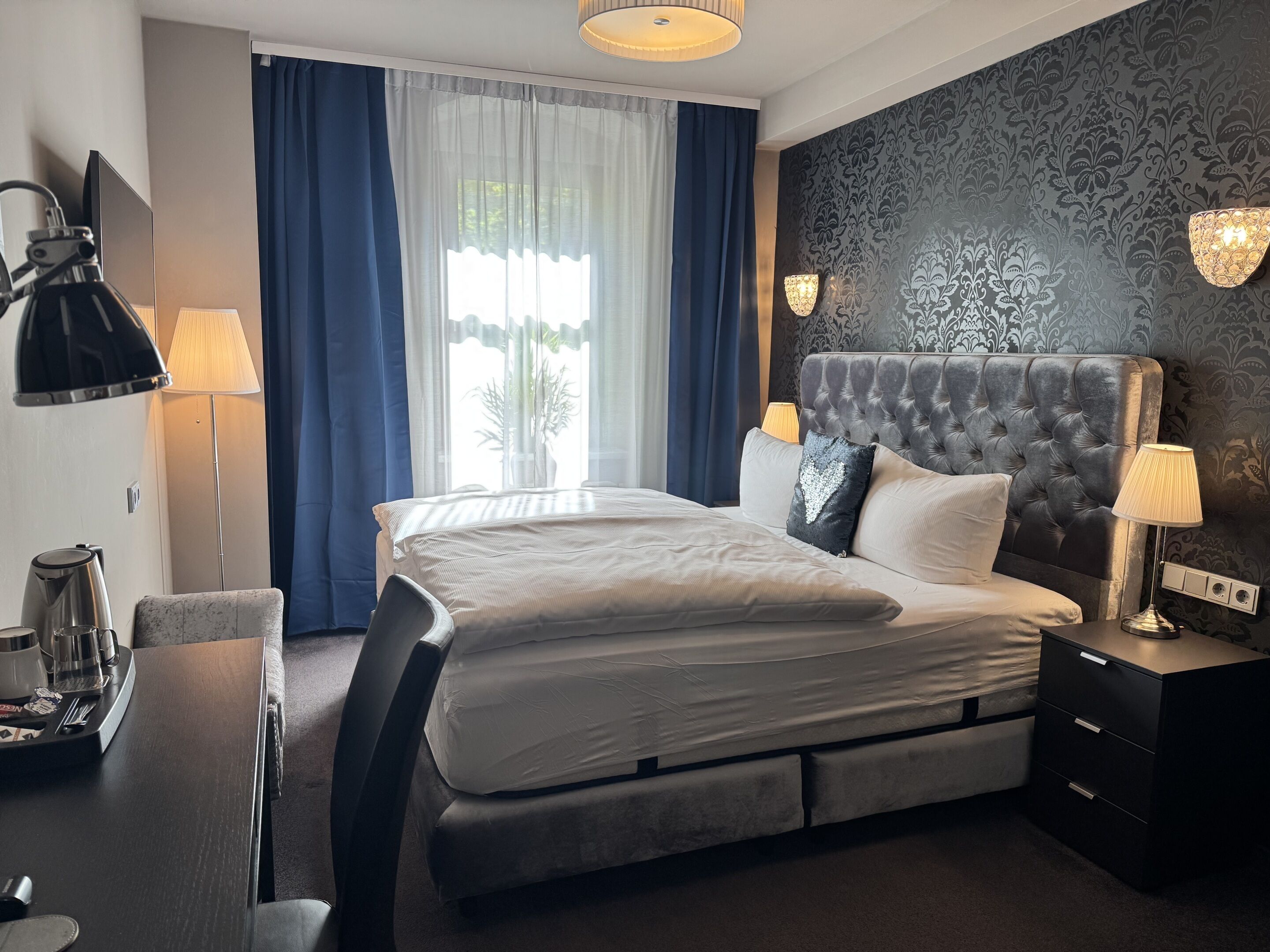 A hotel room with a large bed, a desk with a kettle, a chair, and a lamp.