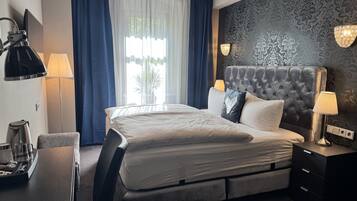 A hotel room with a large bed, a desk with a kettle, a chair, and a lamp.