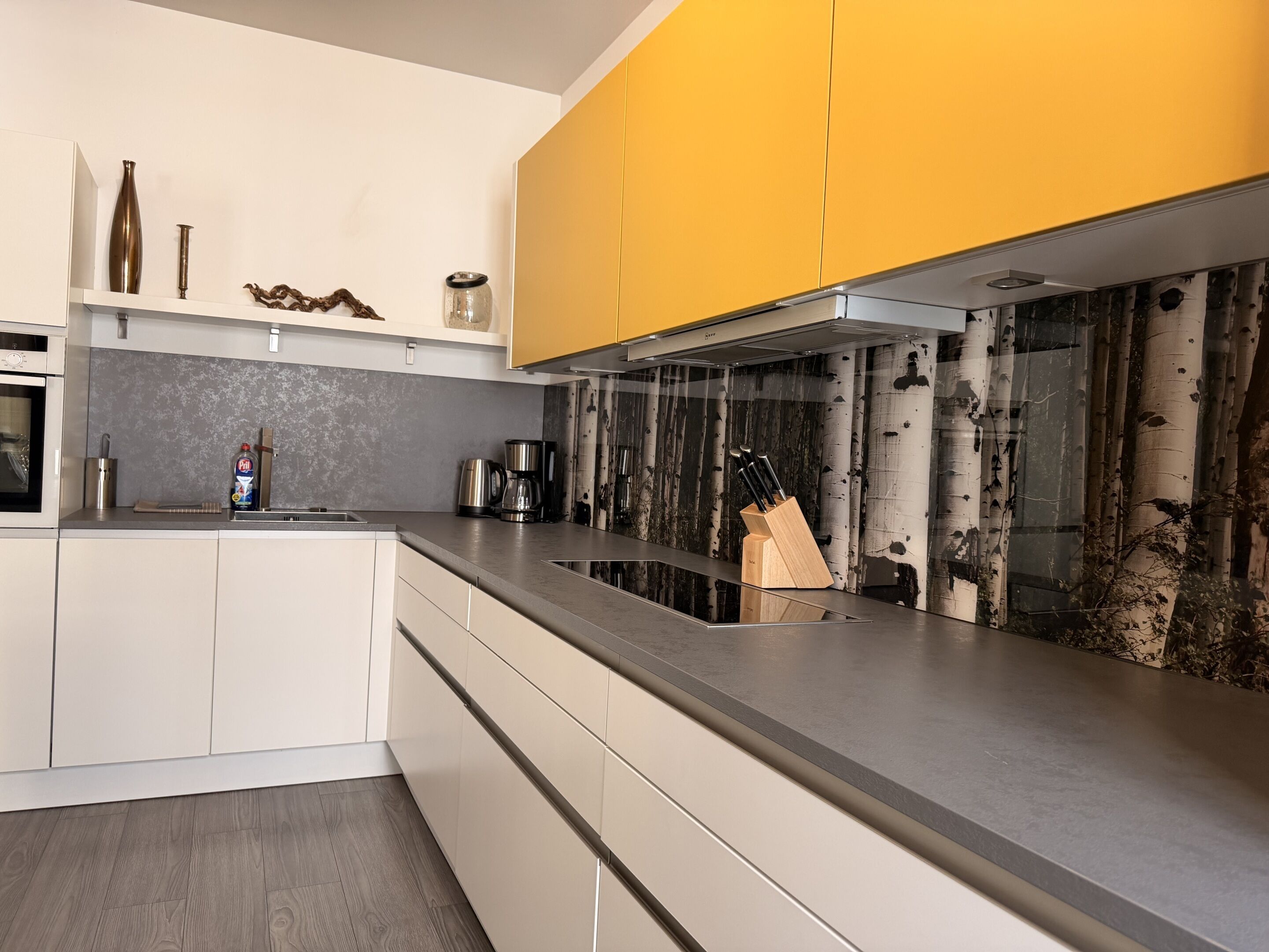 Een moderne keuken met witte kasten, een gele accentmuur, een ingebouwde kookplaat en een messenblok op het aanrecht.