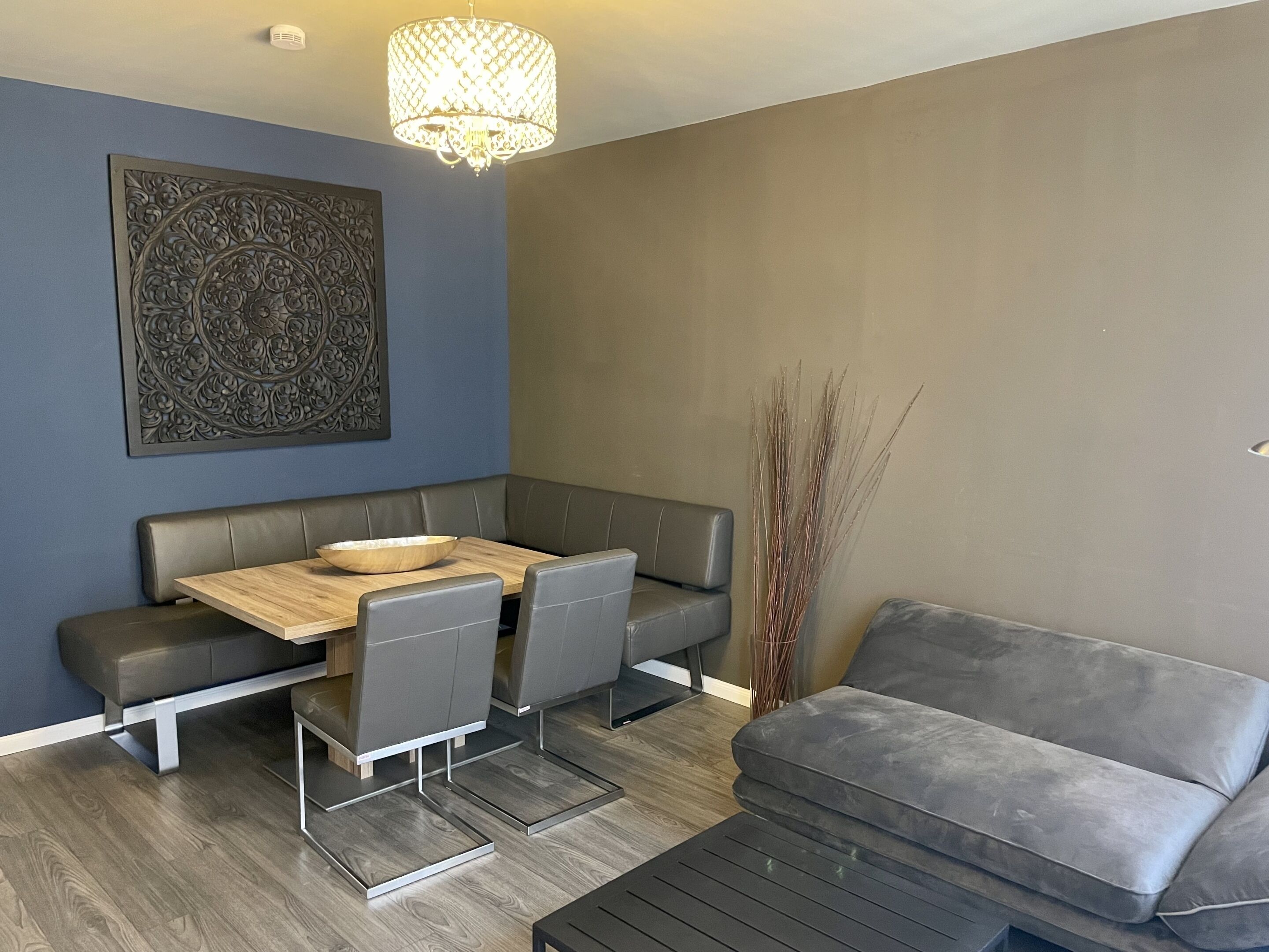Un salon moderne avec un canapé gris, une table à manger avec des chaises, un élément décoratif mural et une grande plante.