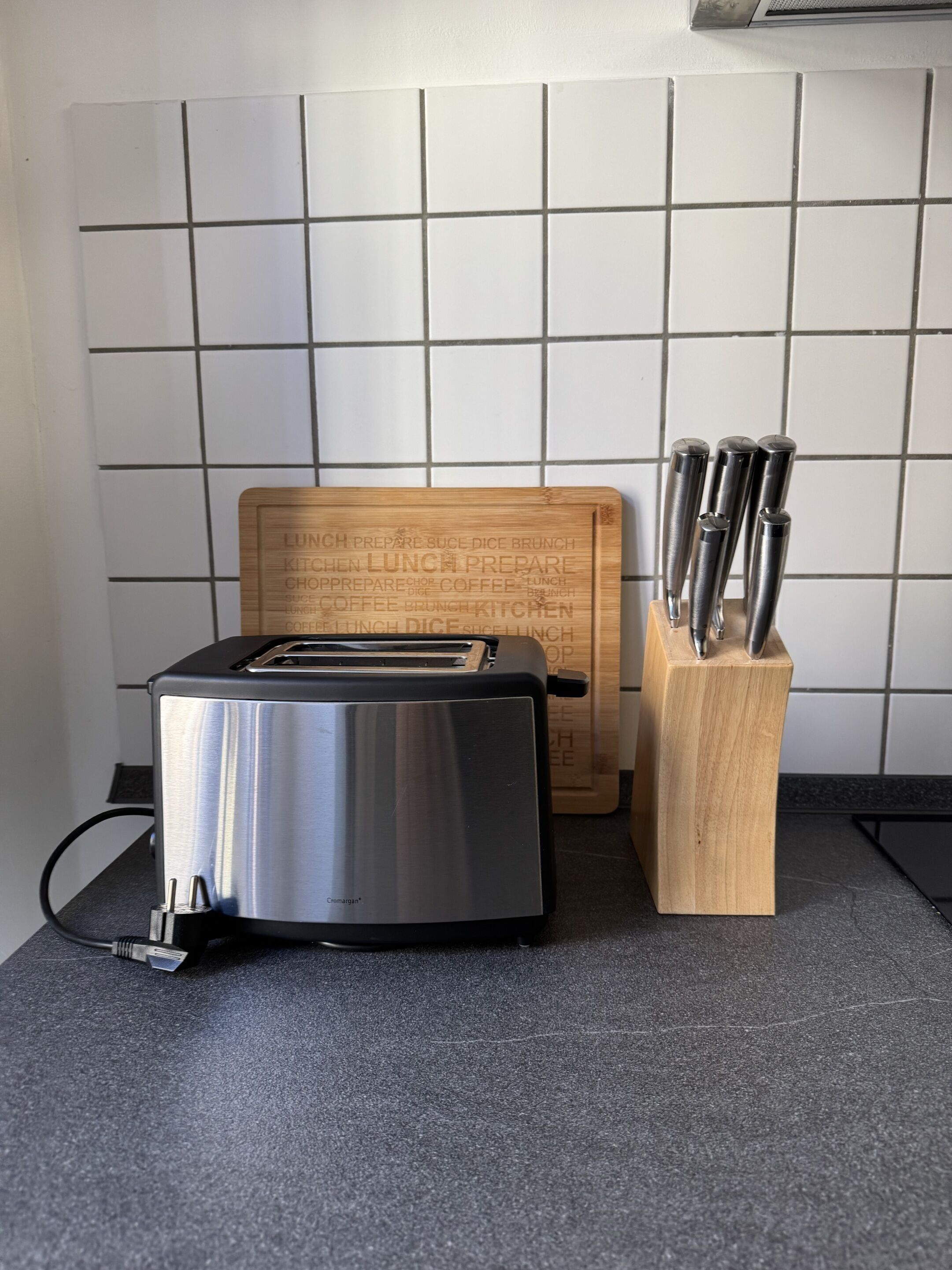 A kitchen with a toaster, a wooden cutting board with kitchen-related words, and a knife block.