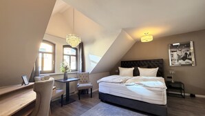A bedroom with a large bed, a desk, a chair, a small table with a vase, and a chandelier. - Pension Burkhardt (Meissen)