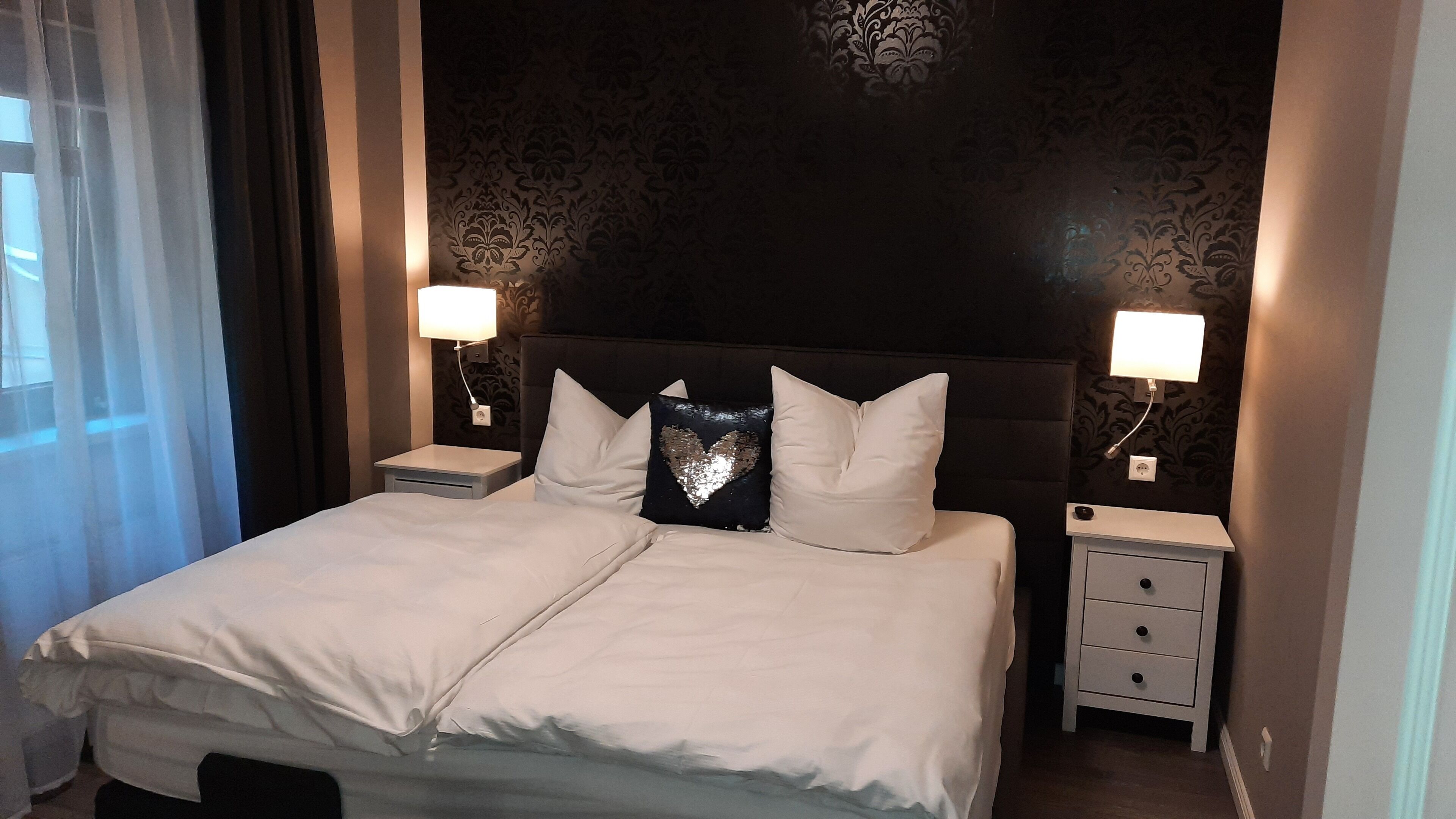 A neatly made bed with white linens, two bedside tables with lamps, and a decorative heart pillow.