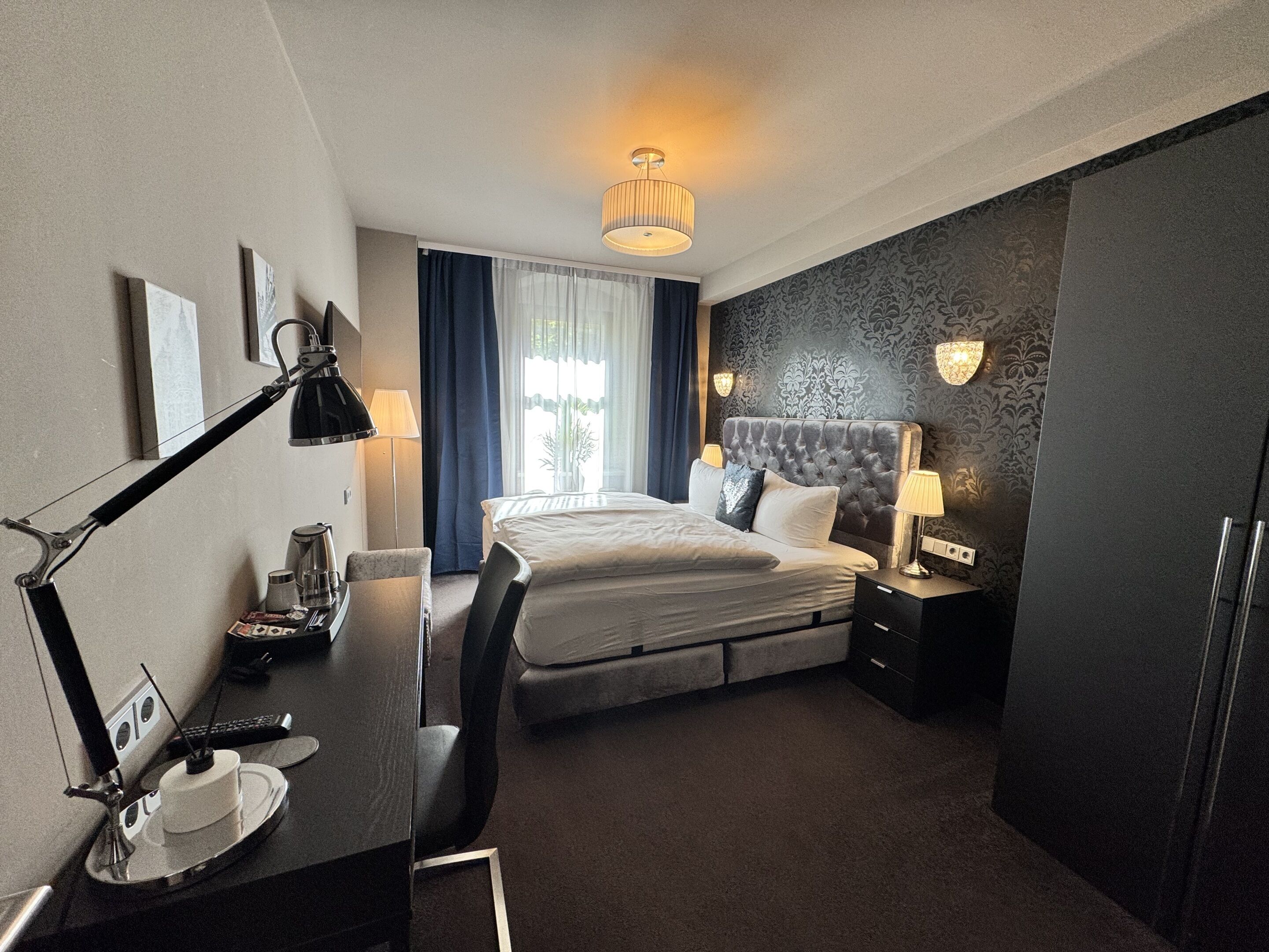 Habitación de hotel con cama, mesitas de noche, escritorio con lámpara, silla y ventana con cortinas.