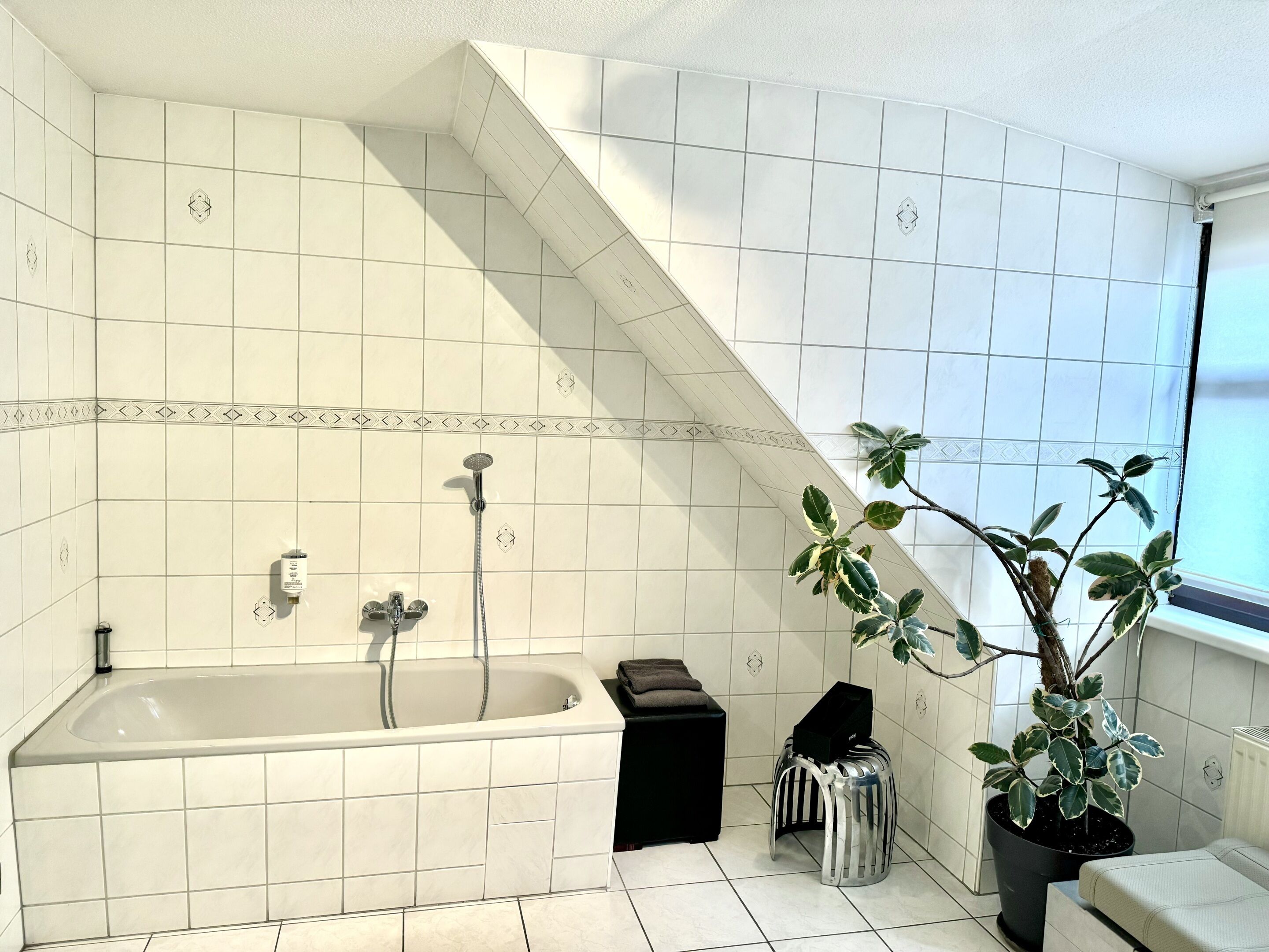 Une salle de bain moderne avec une baignoire sur pieds, un petit tabouret, une plante en pot et une fenêtre laissant entrer la lumière naturelle.