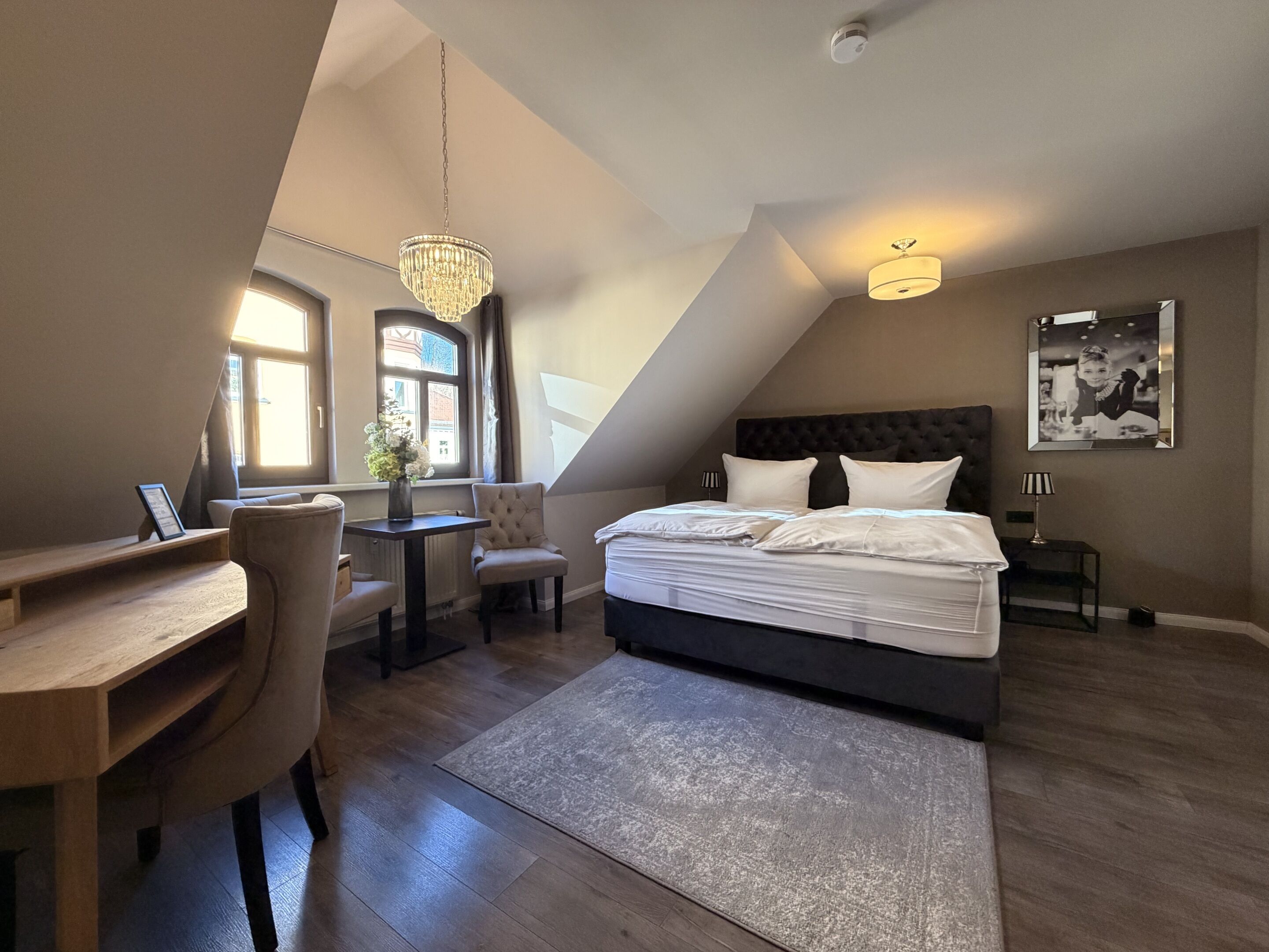 A bedroom with a bed, a desk, a chair, a small table, a chandelier, a lamp, and a framed picture on the wall.