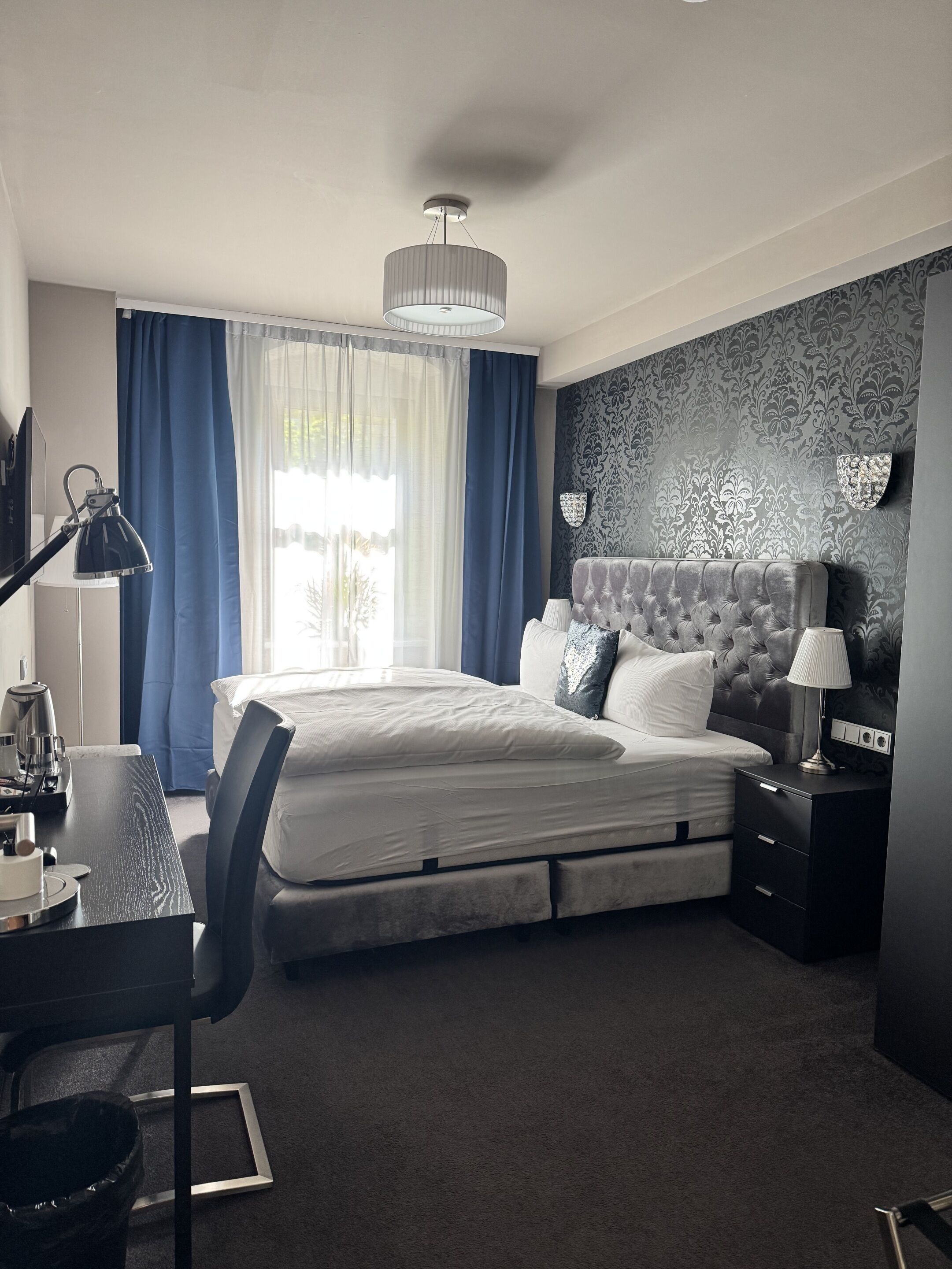 Una habitación de hotel moderna con una cama grande, un escritorio con lámpara, una silla, una mesita pequeña y una mesita de noche con lámpara.