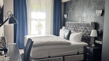 Una habitación de hotel moderna con una cama grande, un escritorio con lámpara, una silla, una mesita pequeña y una mesita de noche con lámpara.