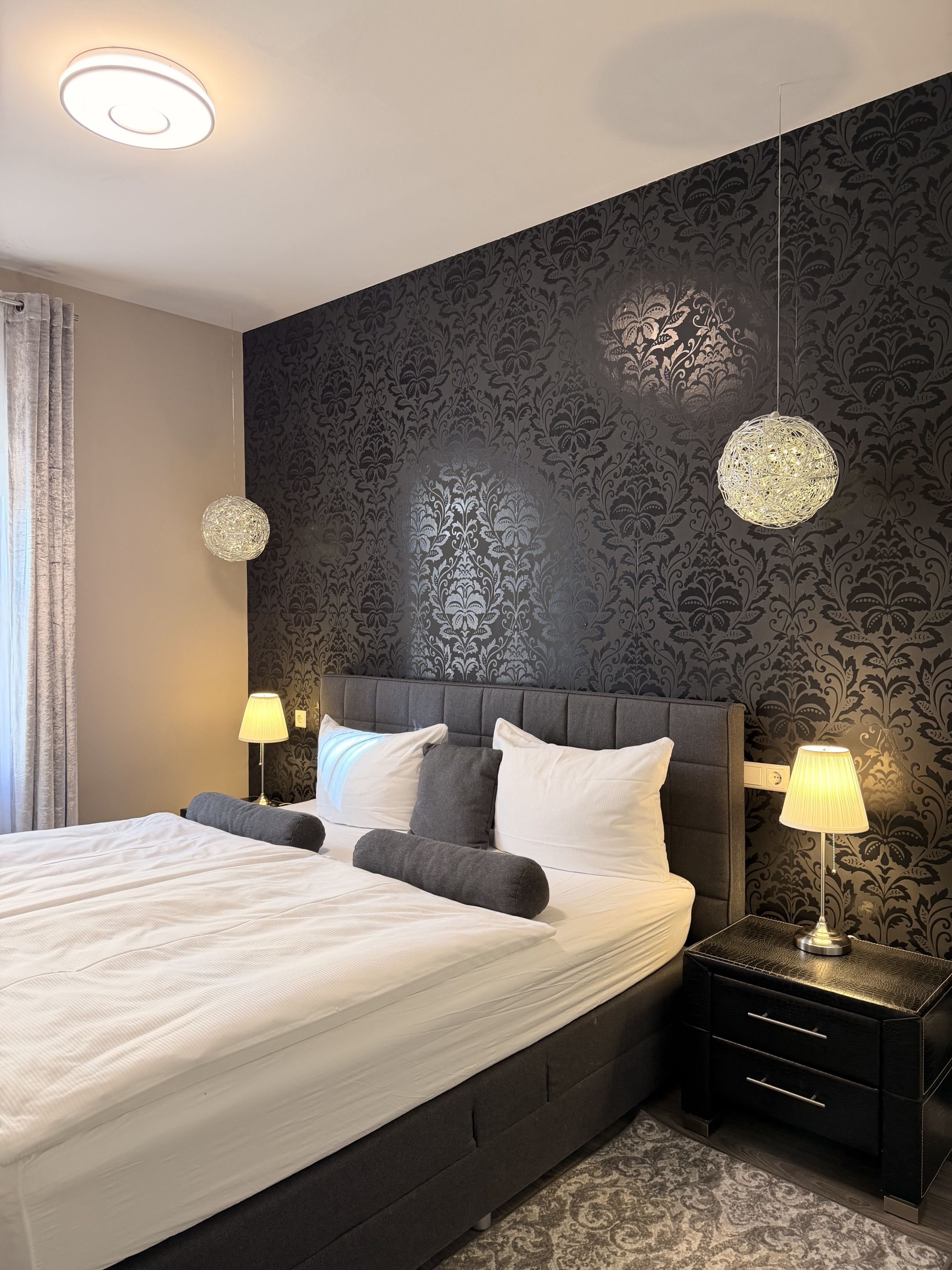 A modern hotel room with a large bed, two bedside tables with lamps, a decorative wall, and a ceiling light.