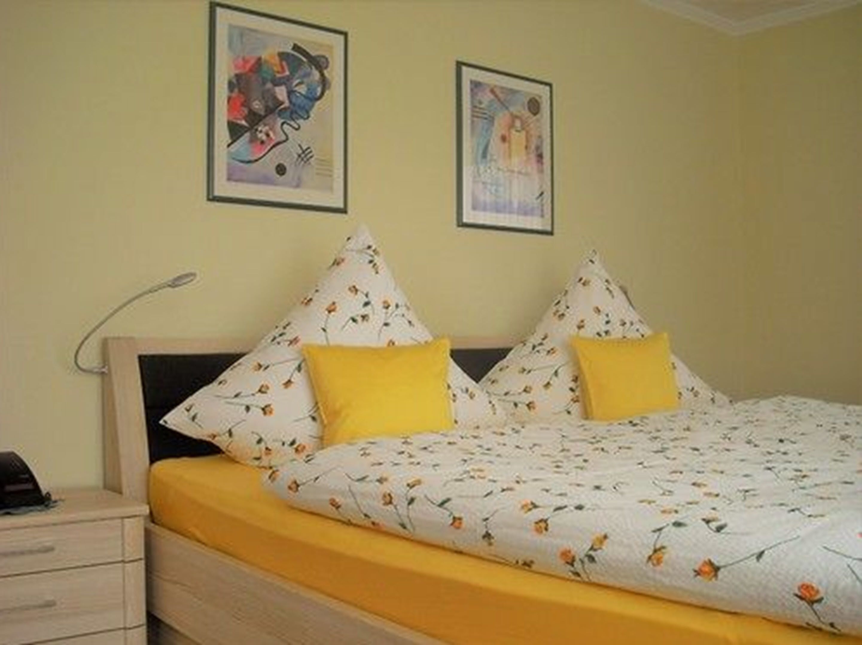 Un dormitorio con cama, dos almohadas, una mesita de noche y dos cuadros enmarcados en la pared.