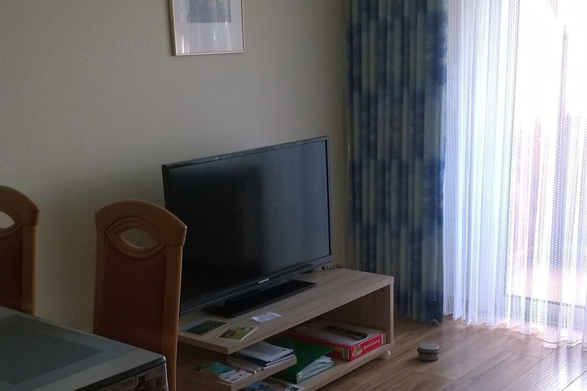 Ein Wohnzimmer mit Fernseher auf einem Holzboden, einem Esstisch mit Stühlen und einem Fenster mit transparenten Vorhängen.