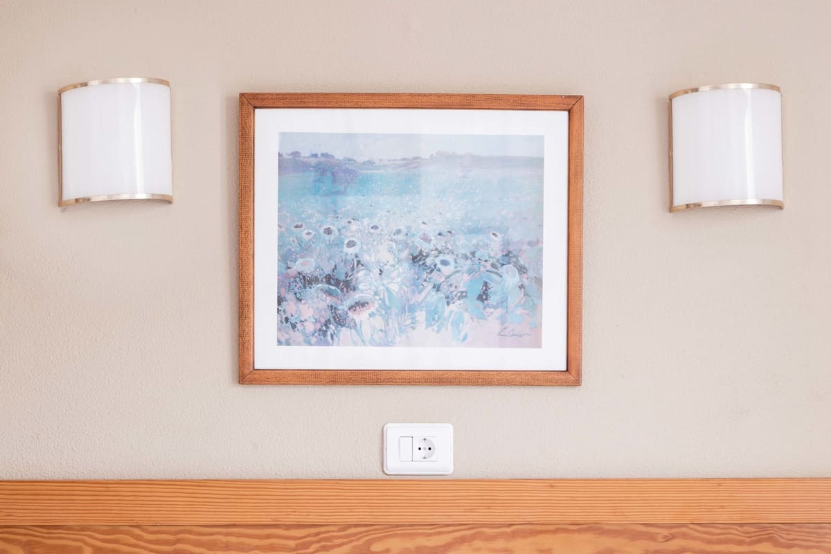 A hotel room with a wooden headboard, a framed abstract painting, and two wall-mounted lamps.