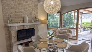 A living room with a stone fireplace, a glass coffee table surrounded by chairs, and a large window offering a view of greenery. - Eco Pebble Bay Beach House (Vela Luka)