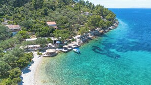 An aerial view of a coastal area with clear turquoise waters, a small pier with a boat, and several buildings surrounded by trees. - Eco Pebble Bay Beach House (Vela Luka)