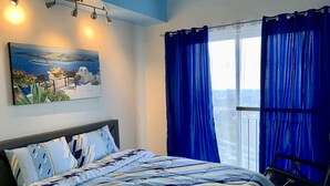 Un dormitorio con una cama, un cuadro y vistas por una ventana con cortinas azules.