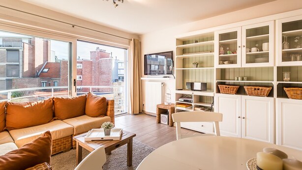 A modern living room with a sofa, a dining table, and a kitchen area with cabinets and a television. - Gaston Lejeune 304 (Koksijde)