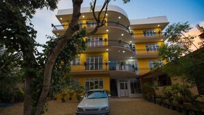 Un edificio de varios pisos con balcones, un coche estacionado adelante y macetas con plantas alrededor.