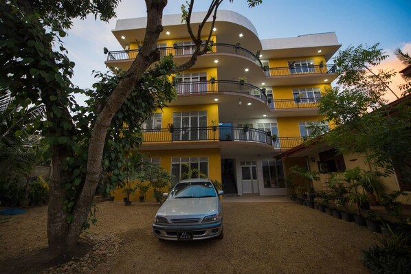 Un edificio de varios pisos con balcones, un coche estacionado adelante y macetas con plantas alrededor.
