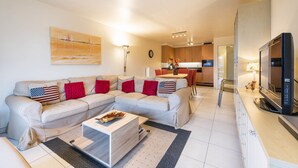 A modern living room with a sectional sofa, a coffee table, and a TV. There is a kitchen area in the background with a dining table and chairs. - Pablo II-301 (Koksijde)