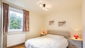 A bedroom with a large bed, a window with curtains, a bedside table with a lamp, and a radiator. - Pablo II-301 (Koksijde)