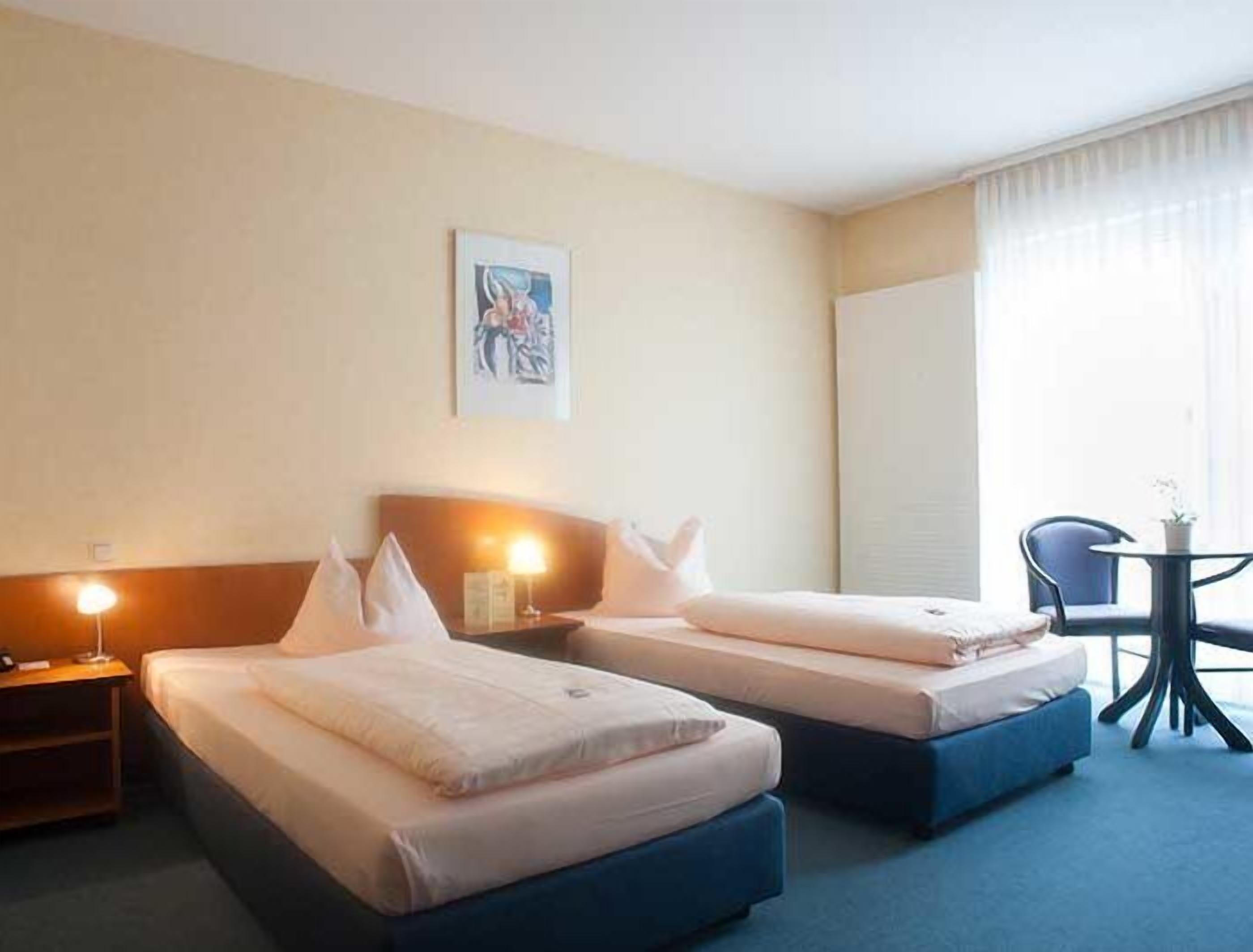 A hotel room with two beds, a small table with a chair, and a painting on the wall.