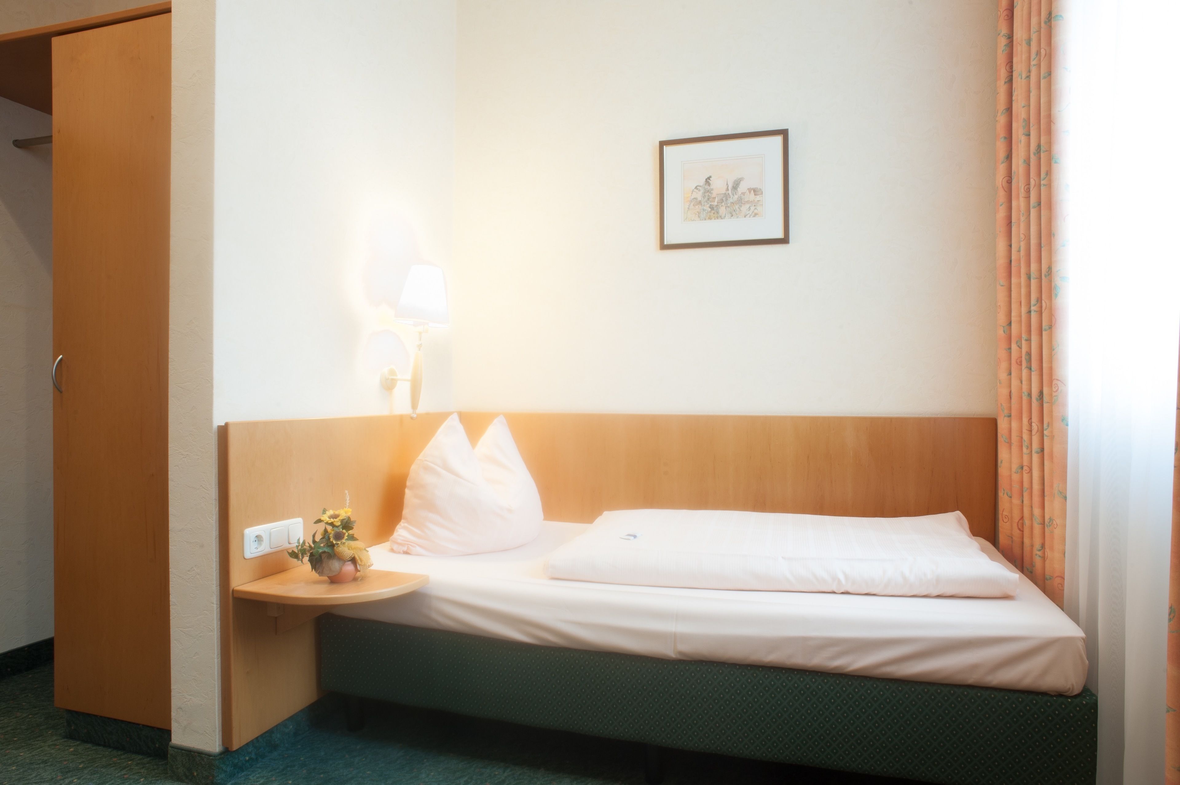 A hotel room with a bed, a bedside table with a lamp, a framed picture on the wall, and a window with curtains.