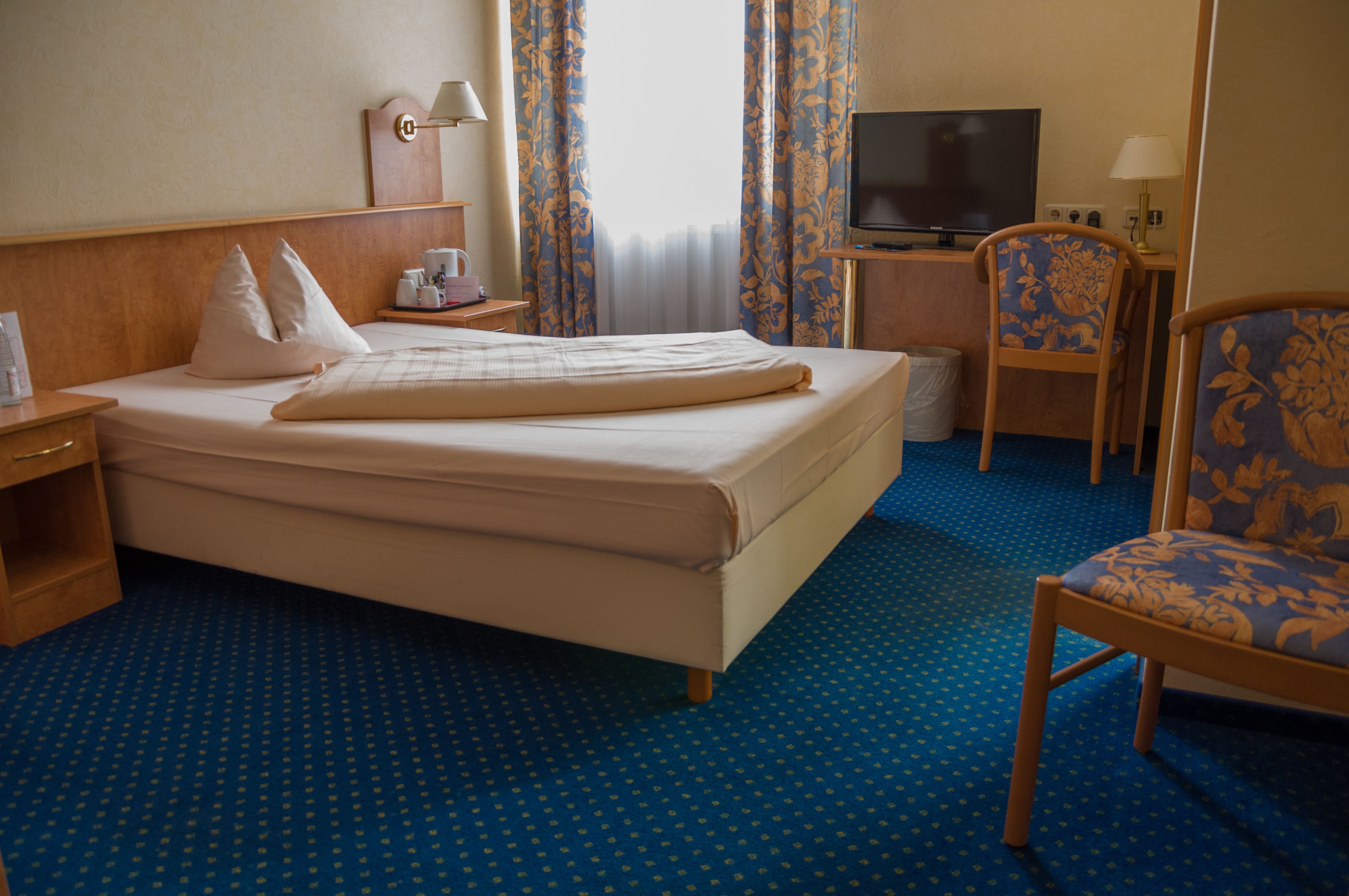 Habitación de hotel con cama, escritorio con televisor, una silla y mesita de noche con lámpara.