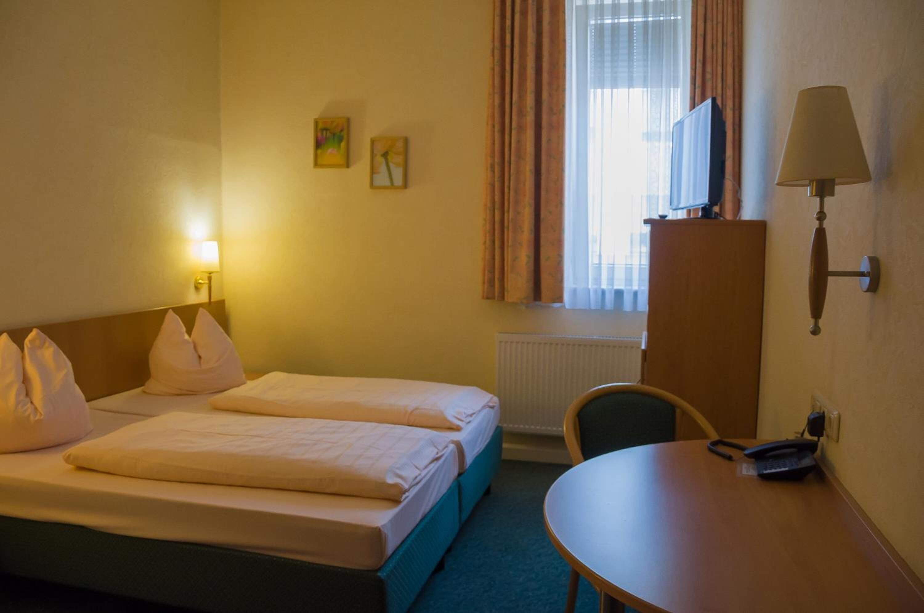 A hotel room with two beds, a desk, a chair, a TV, and a lamp.
