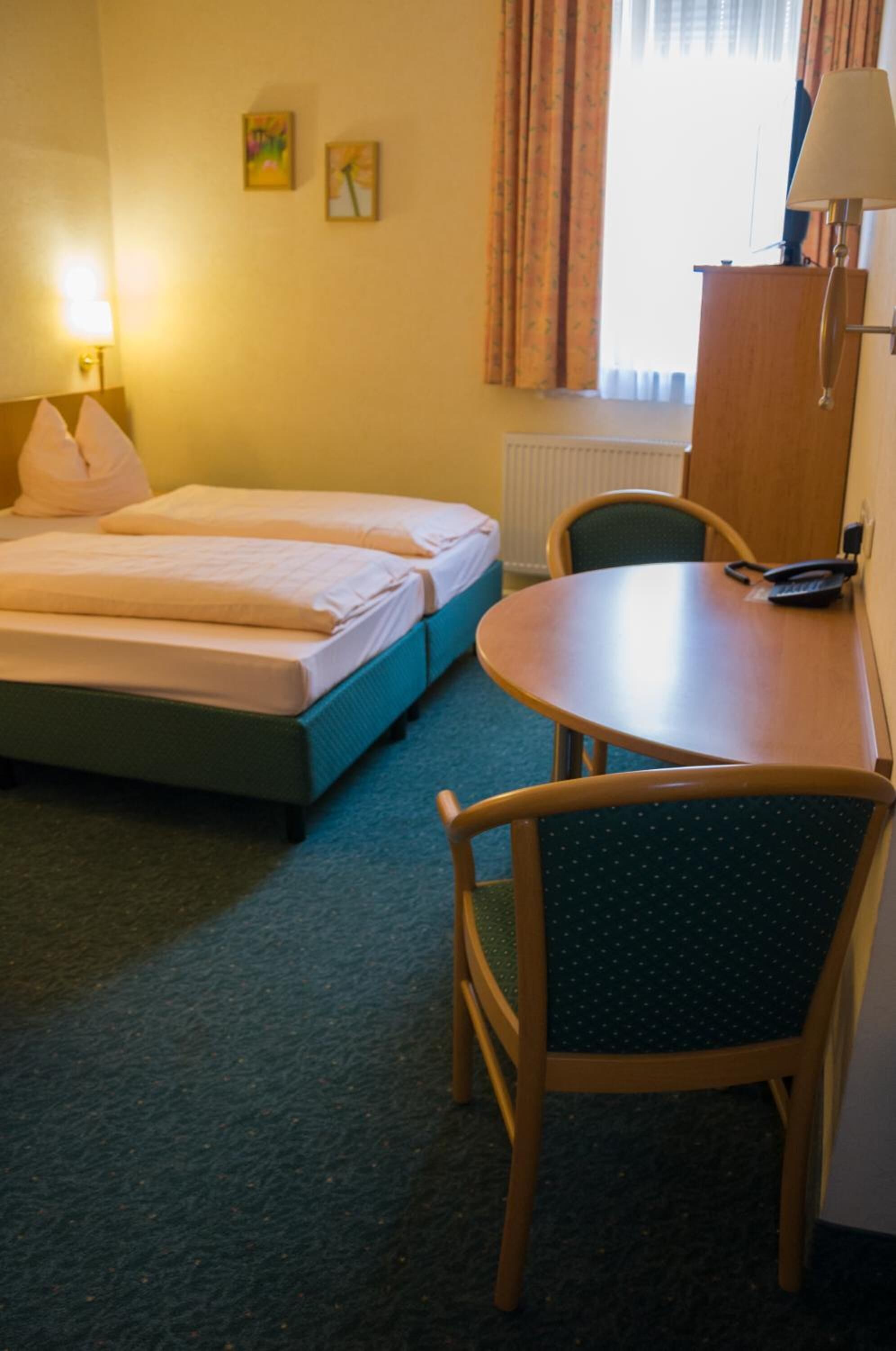 Ett hotellrum med två sängar, ett skrivbord, en stol, en lampa och ett fönster med gardiner.