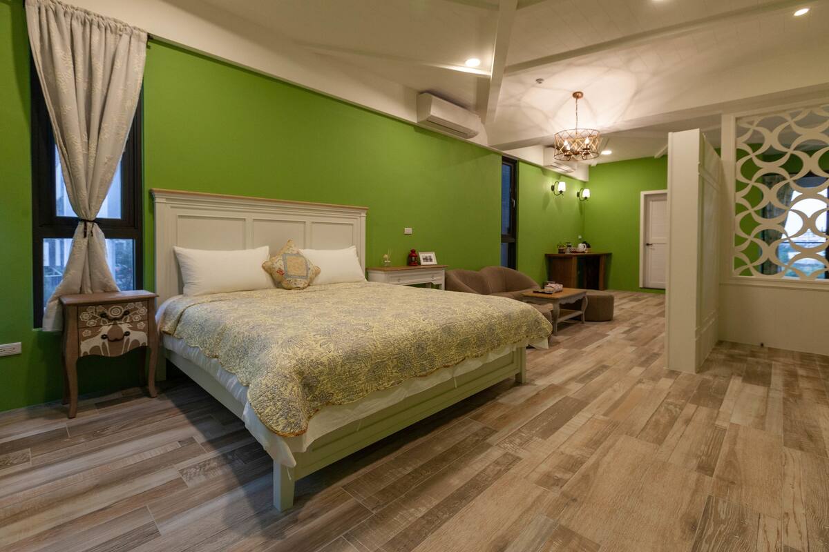 A bedroom with a green wall, a wooden floor, a bed with a white headboard, a nightstand, a chair, a table, and a window with a curtain.