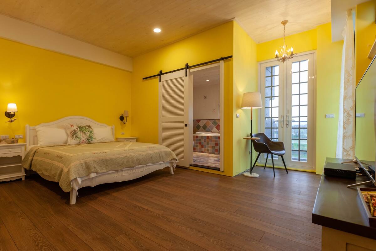 A bedroom with a bed, bedside tables, a lamp, a chair, a desk with a computer, and a sliding door leading to a bathroom.
