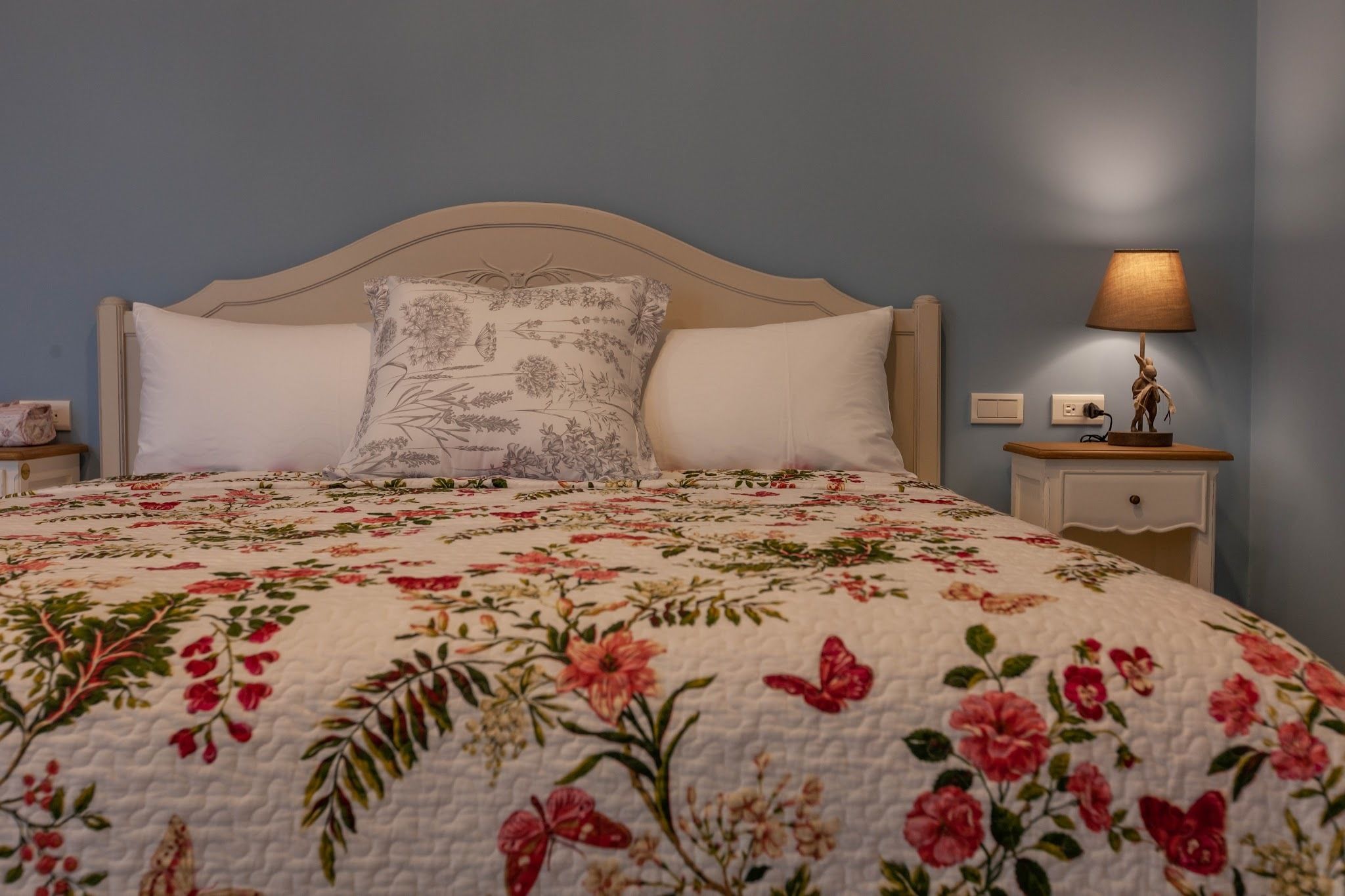 Ein Schlafzimmer mit einer floralen Steppdecke, einem Holzkopfende, einer Nachttischlampe und einem Nachttisch.