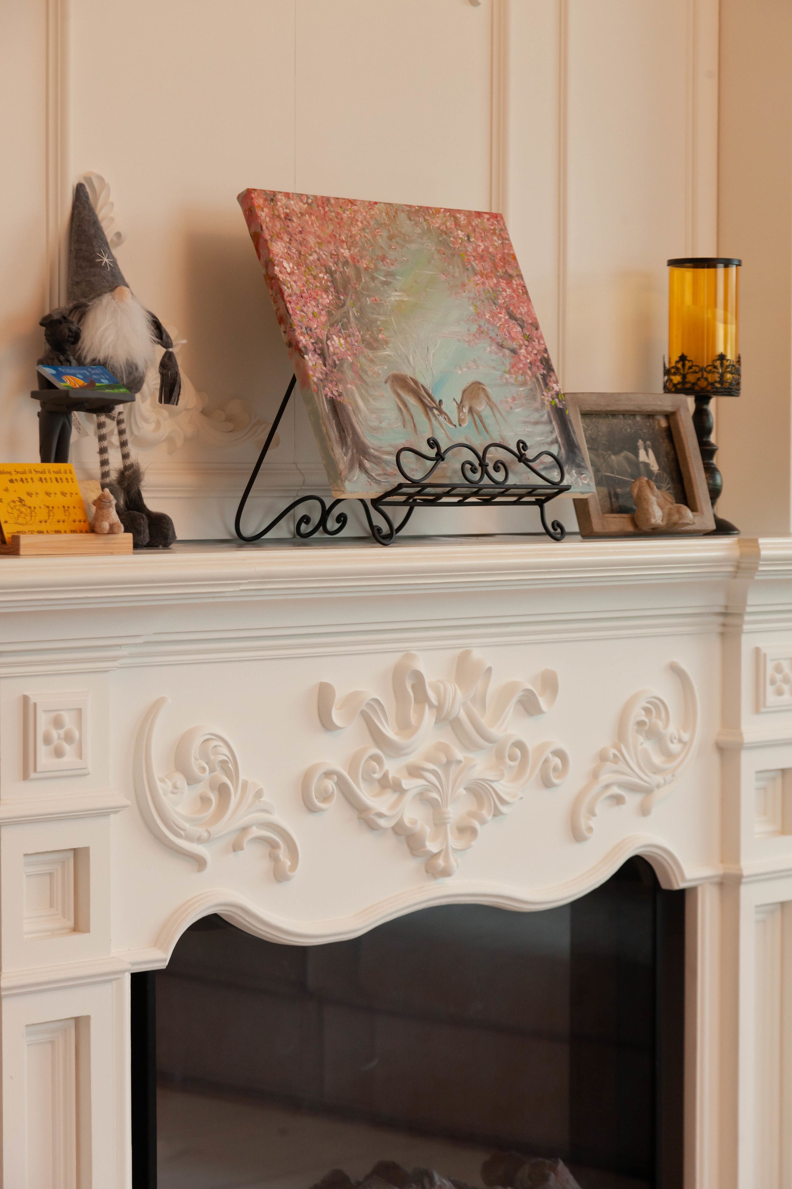 A fireplace with ornate white mantel, a framed painting of a landscape, and a gnome figurine.