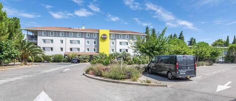 Un hotel de varios pisos con fachada amarilla y blanca, un estacionamiento con una furgoneta y un cielo azul despejado.