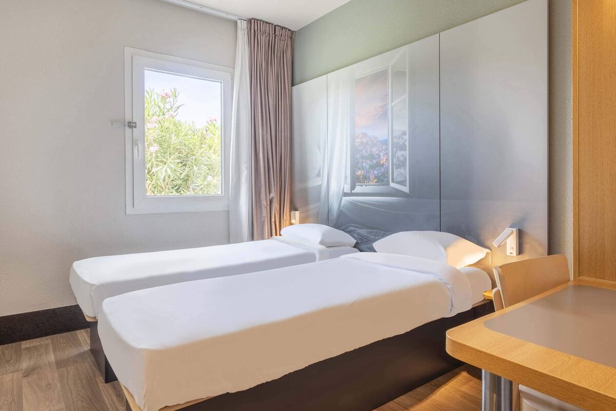Habitación de hotel con dos camas, una ventana con cortinas, una mesita de noche y vistas a zonas verdes.