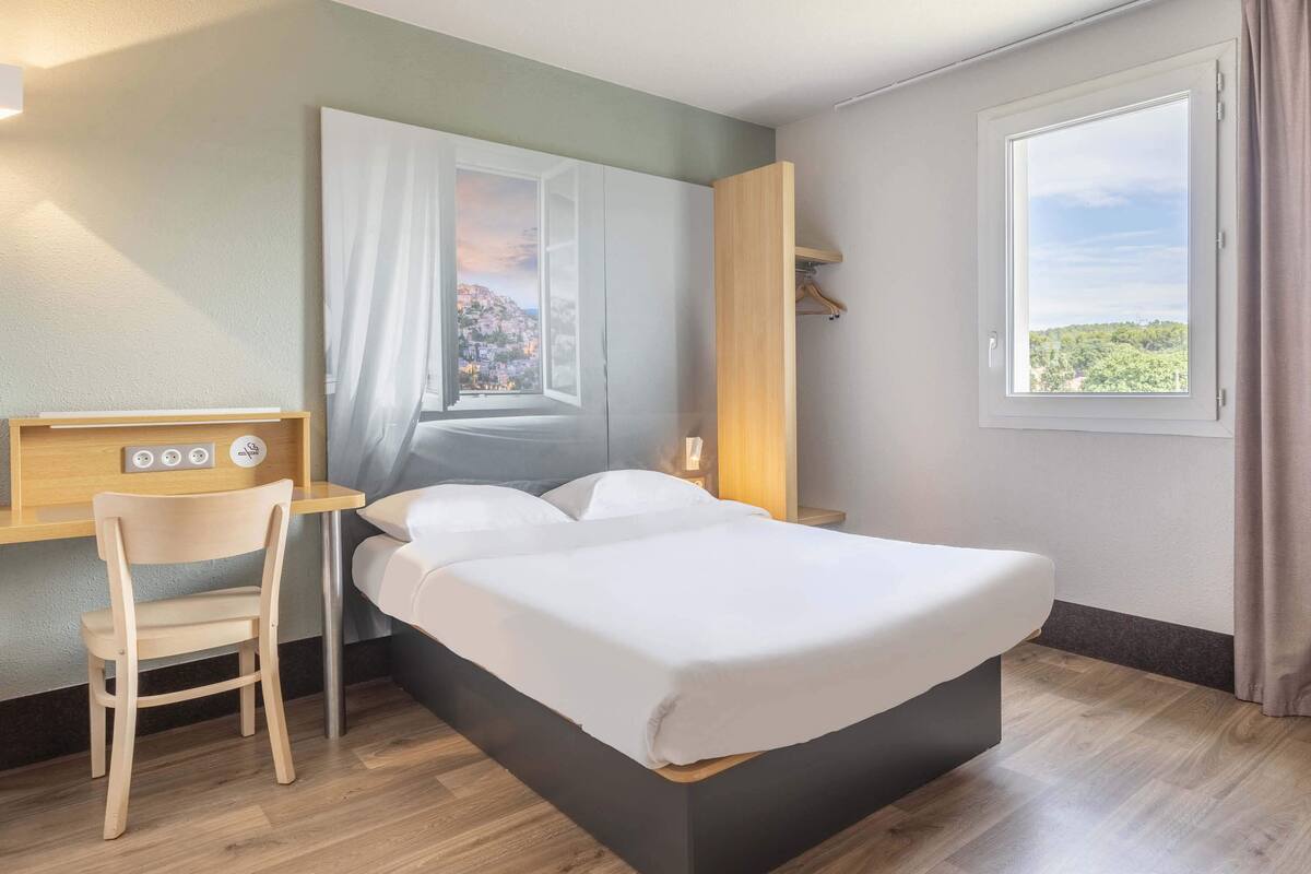 Habitación de hotel con cama, escritorio con silla, ventana con vistas y estante fijado a la pared.