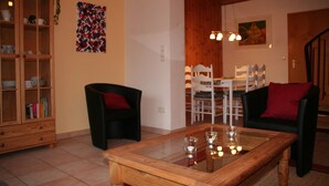 A living room with a glass-top coffee table, two black armchairs, a wooden cabinet, and a dining area with white chairs and a table.
