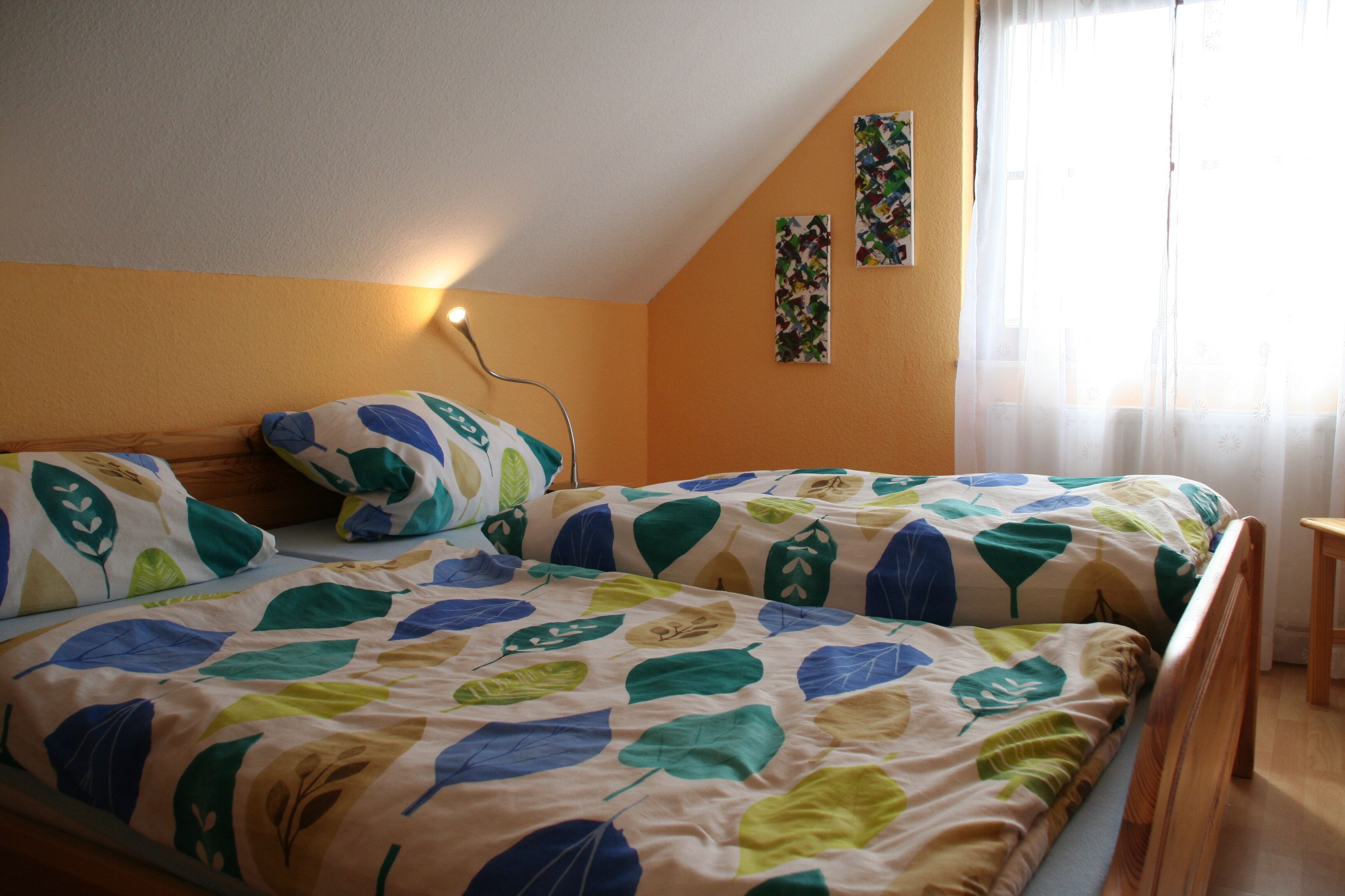 Une chambre avec deux lits, une tête de lit en bois, une lampe fixée au mur et une fenêtre ornée de rideaux légers.
