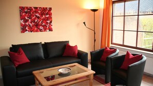A living room with a black sofa, two armchairs, a glass coffee table, a red abstract painting, a floor lamp, and a window with curtains.