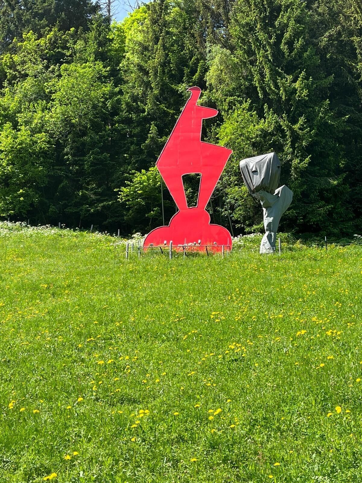 A red abstract sculpture in a grassy field with trees in the background.