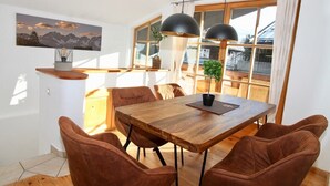 A dining area with a wooden table and brown chairs, a painting on the wall, and large windows with curtains.