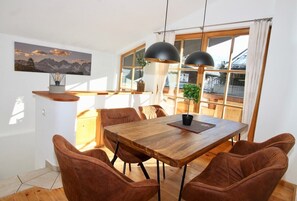A dining area with a wooden table and brown chairs, a painting on the wall, and large windows with curtains.
