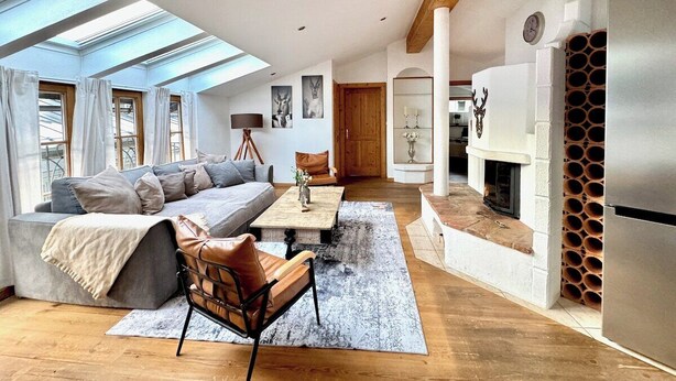 A modern living room with a sofa, coffee table, and leather chair. There is a fireplace with a log storage, a skylight, and a large window.