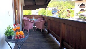 A wooden balcony with wicker chairs, a small table with fruit and drinks, and a view of houses and mountains.