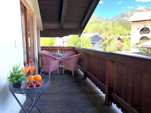 A wooden balcony with wicker chairs, a small table with fruit and drinks, and a view of houses and mountains.