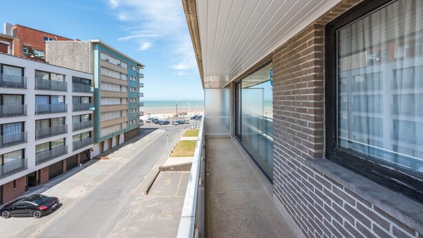 Un balcón con vista a un estacionamiento y edificios, cielo despejado y una playa a lo lejos.