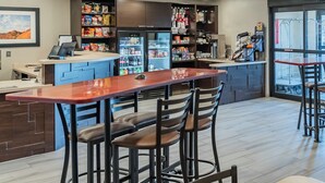 Een moderne bar met een lange toonbank, drie krukken en een tafel. Een koelkast is gevuld met drankjes en snacks, en er staat een koffieapparaat. De ruimte heeft een strak, eigentijds design met een houten vloer en een groot raam dat natuurlijk licht binnenlaat.