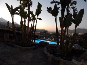 A pool area with lounge chairs, surrounded by tall banana plants, set against a cityscape at dusk. - B&B Sole e Luna (Palermo)