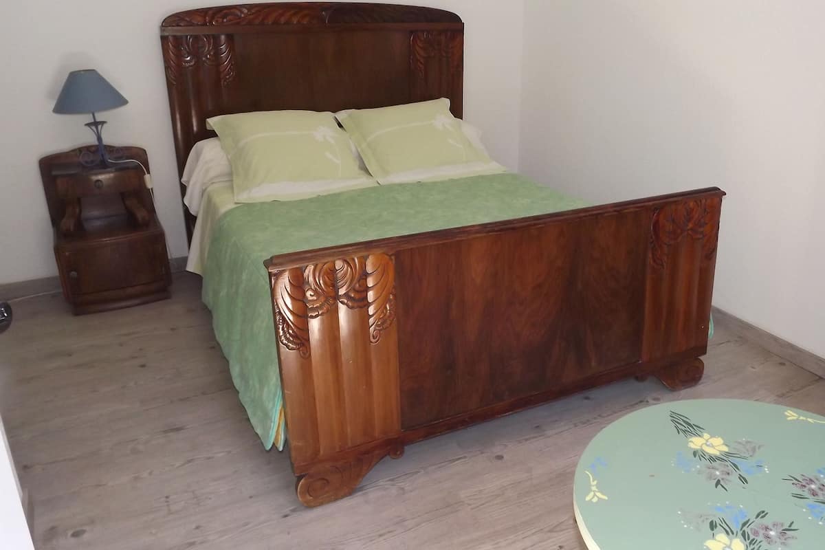 A wooden bed with a green bedspread and pillows, a bedside table with a lamp, and a small round table with floral designs.