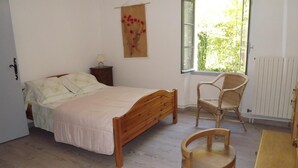 A bedroom with a wooden bed, a wicker chair, a small table, and a window with a view of greenery. - La Bastide B&B (Cavaillon)